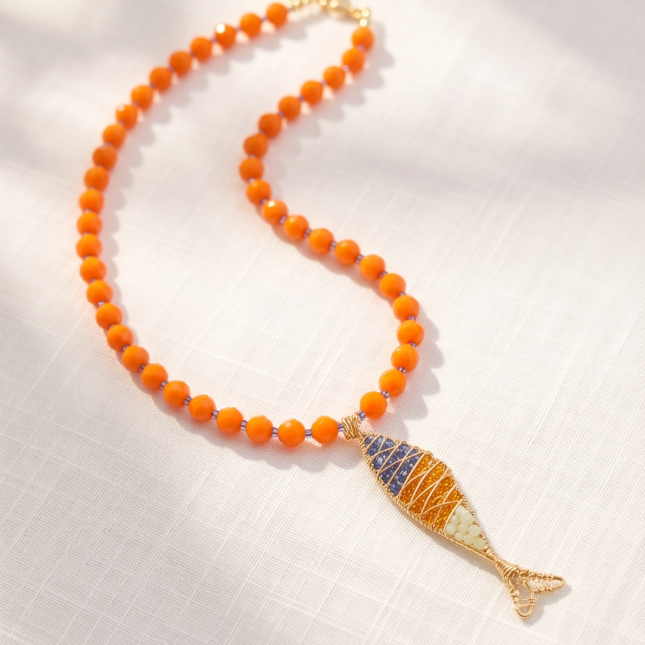 Orange beaded necklace with a gold-tone wire-wrapped fish pendant featuring blue, amber, and cream details on white fabric.
