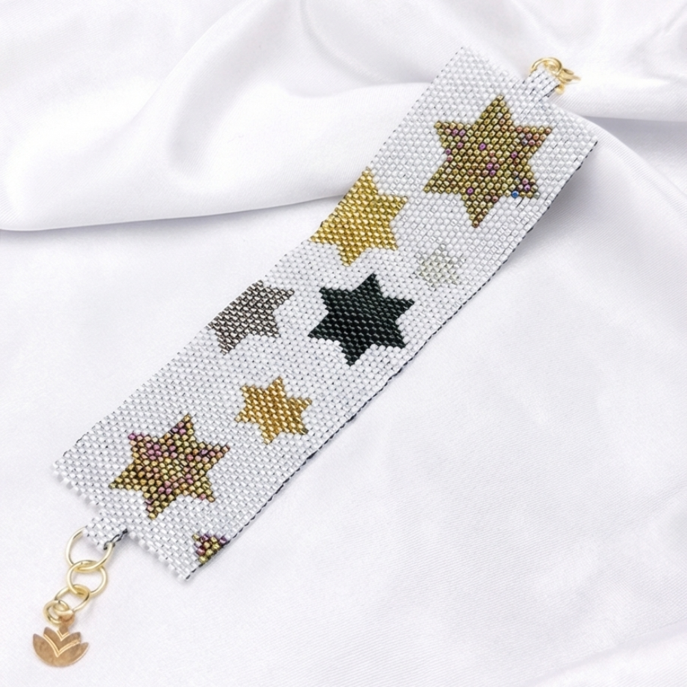 Dailot white beaded bracelet with black and metallic stars, a gold-tone extender chain, and a lotus charm on white fabric.