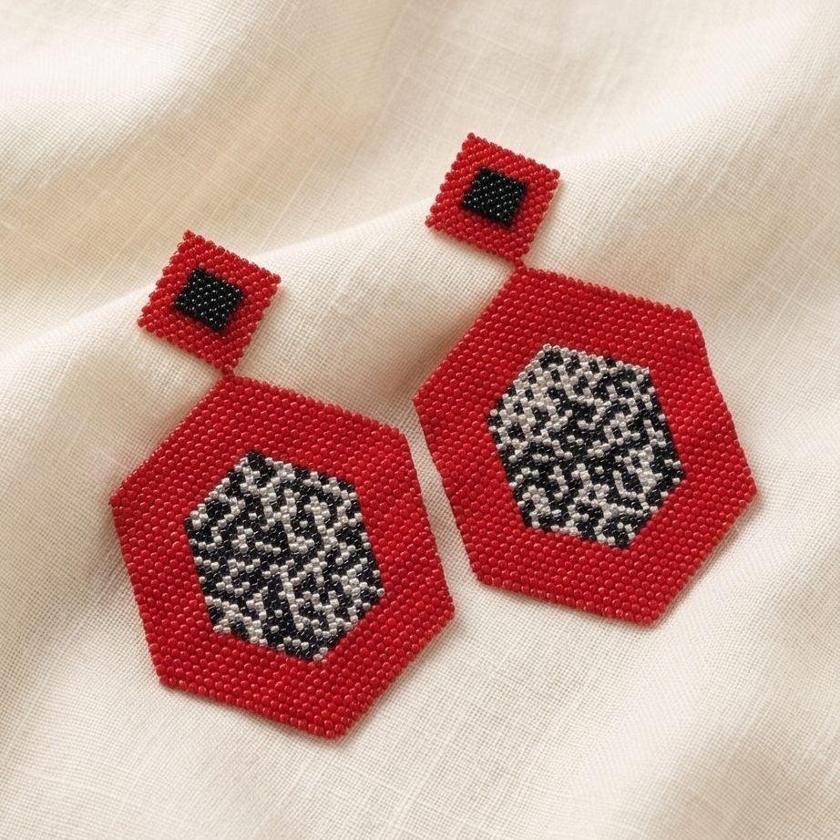 Pair of red hexagonal beaded drop earrings with black and silver-tone patterned centers resting on cream fabric.