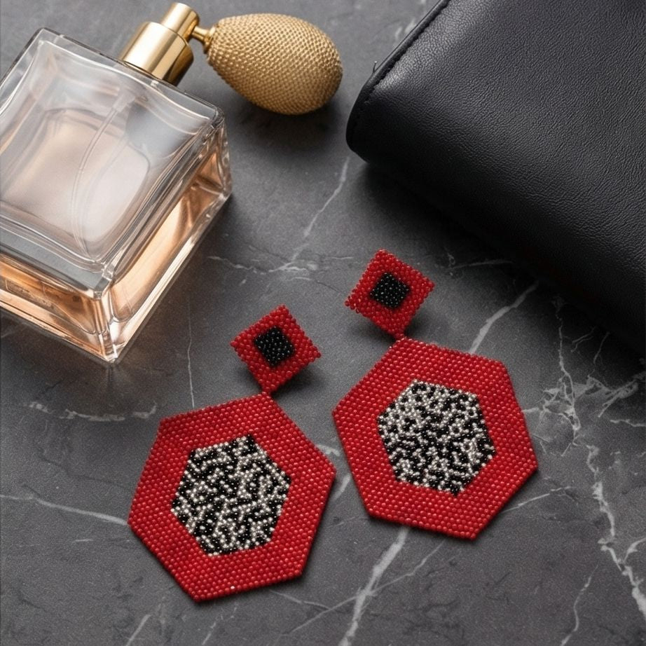 Red geometric beaded earrings with black and silver-tone centers on gray marble beside a perfume bottle and black clutch.