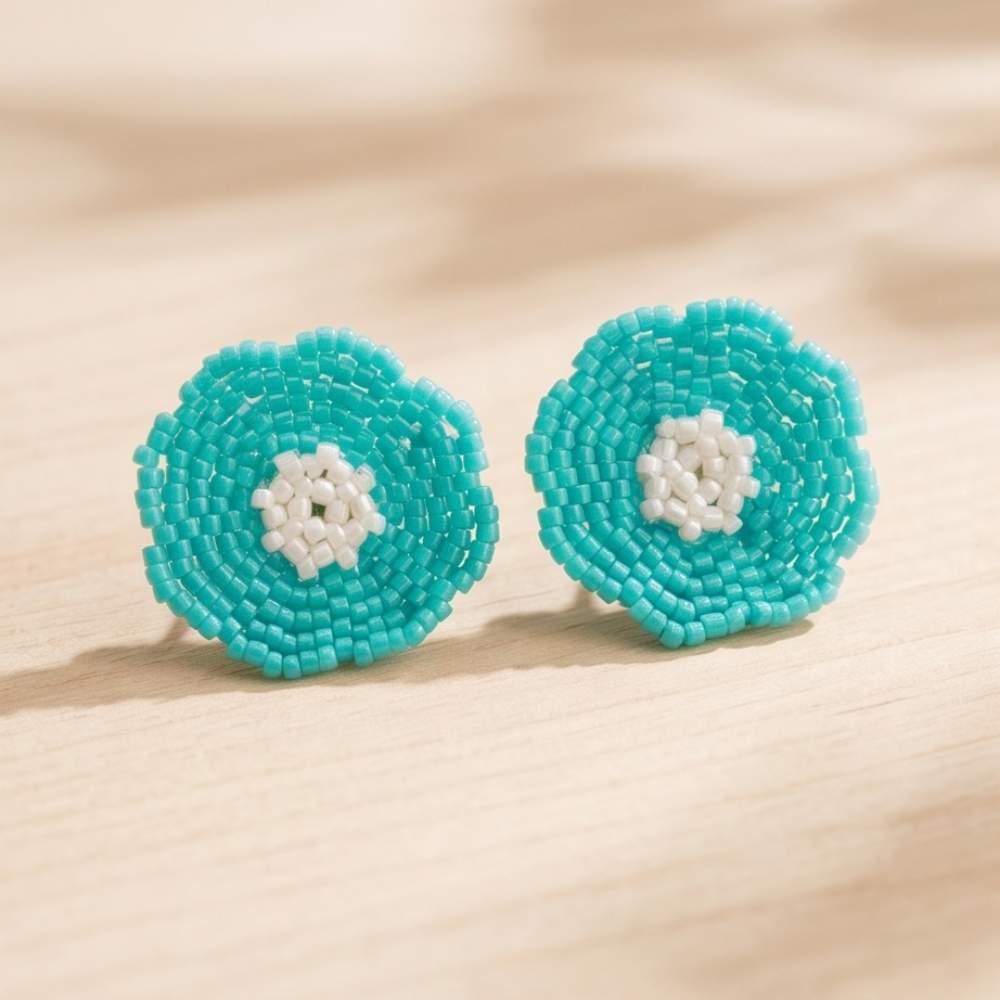 Pair of turquoise beaded flower stud earrings with pearl-white centers on a light wooden surface.