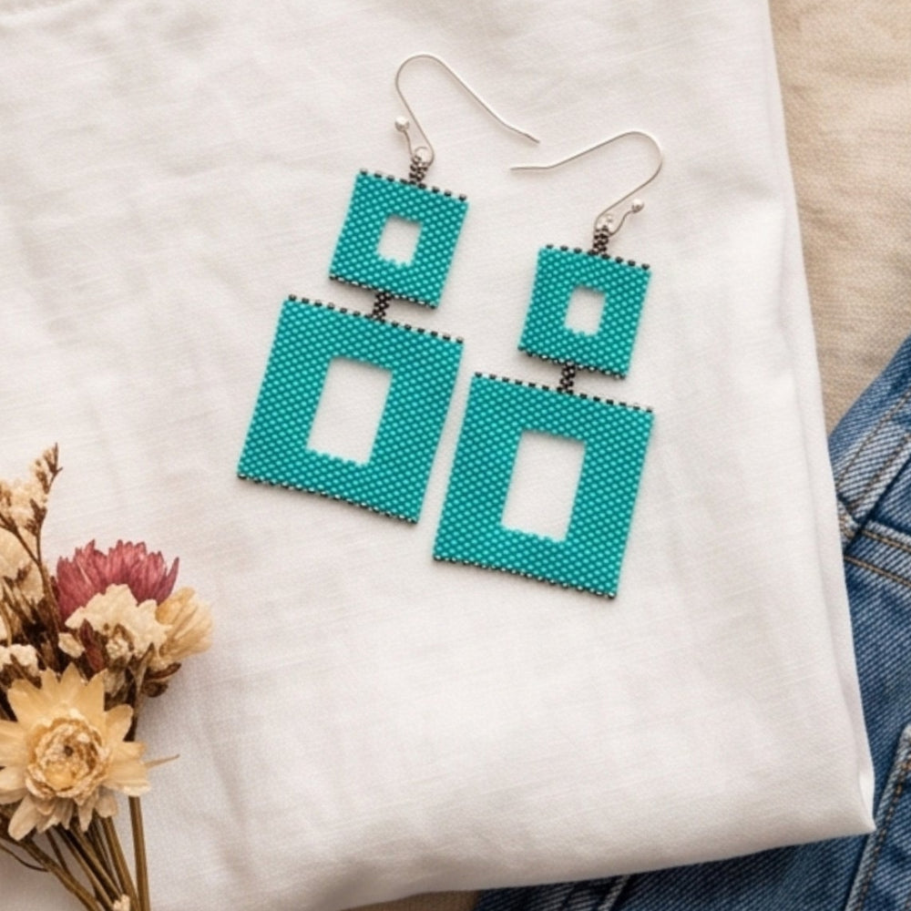 Turquoise double square beaded earrings on a folded white T-shirt beside blue jeans, silver-tone rings, and dried flowers.